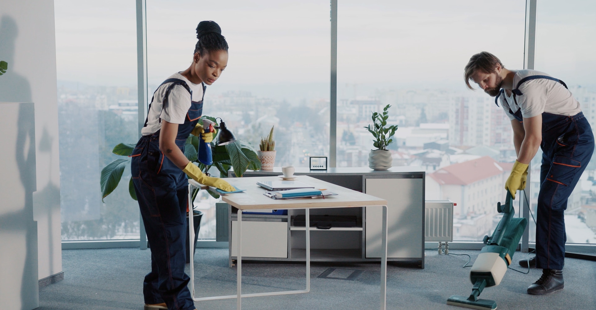 Femme noire et homme blanc, uniformes de nettoyage, nettoyant un bureau moderne avec vue sur la ville et époussetant le dessus d'une table.