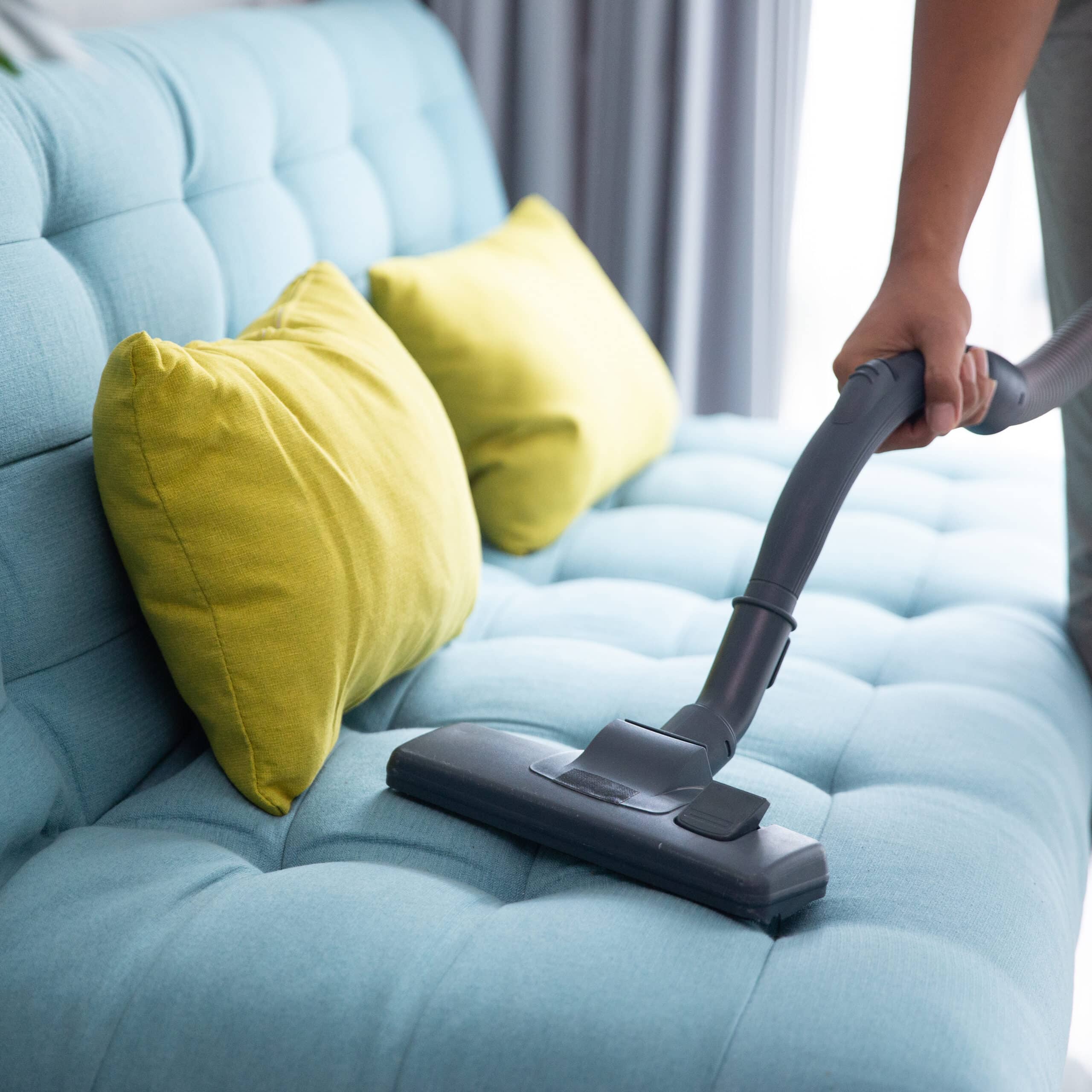 Main tenant un embout d'aspirateur pour nettoyer un canapé bleu clair capitonné avec des coussins jaune vif.